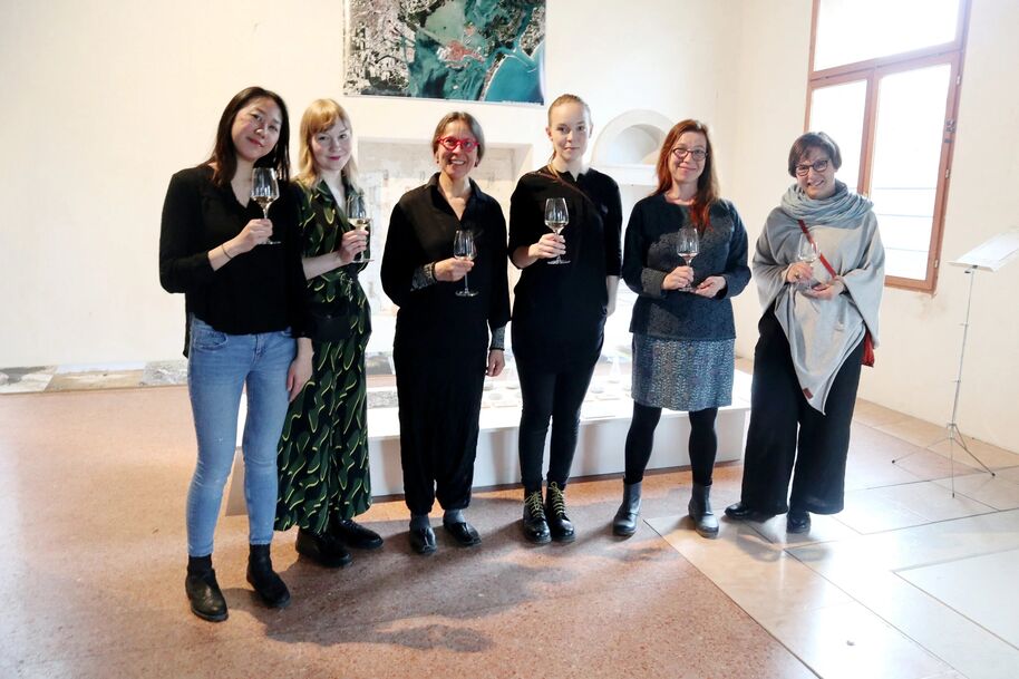 Six people standing indoors holding wine glasses, with artwork on the wall and a large window to the right.