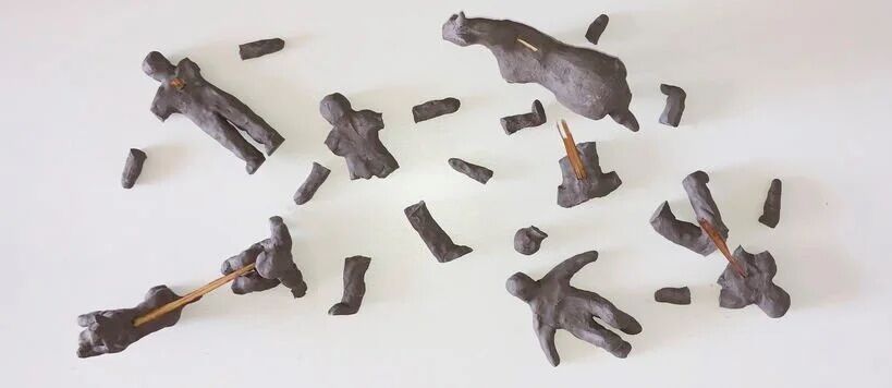 Several small, dark grey clay figures and fragments scattered on a white surface. Some figures have wooden sticks attached.