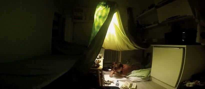 A person lies under a makeshift tent made of blankets, illuminated by a lamp, reading a book in a dimly lit room.