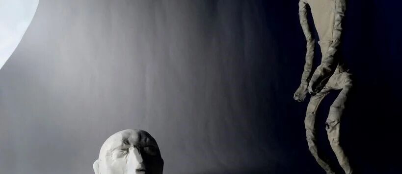 A clay head with closed eyes and a tall, thin figure in the background against a dark grey backdrop.