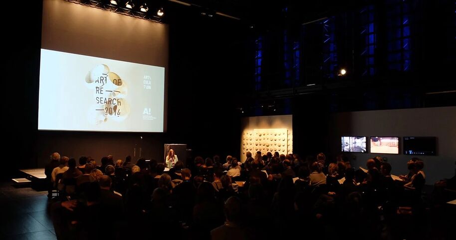 A conference room with attendees seated facing a large screen displaying 'Art of Research 2016'.