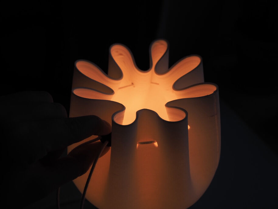A hand adjusts a unique lamp with petal-like openings, emitting a warm glow in a dark room.