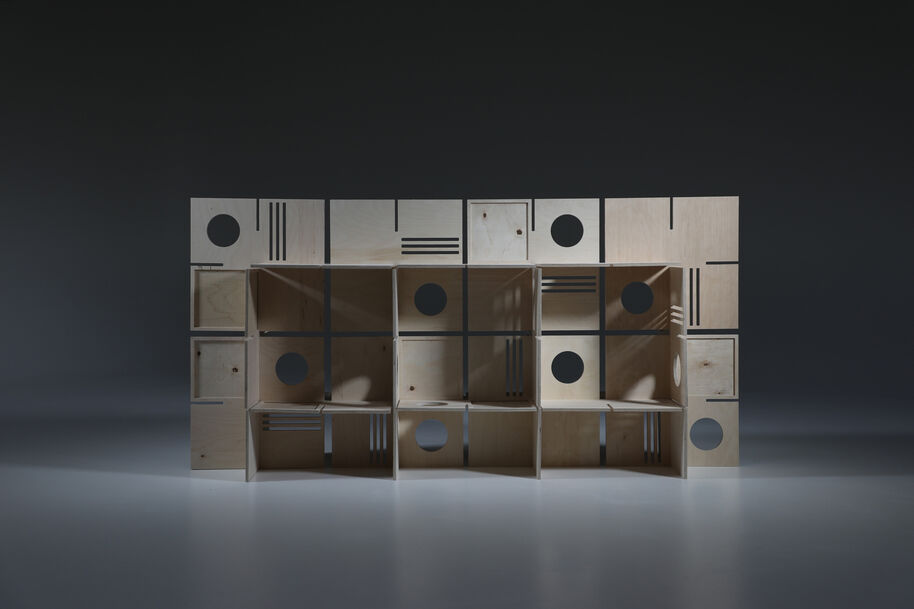 Wooden shelving unit with circular and linear cutouts, arranged in a grid pattern against a dark background.