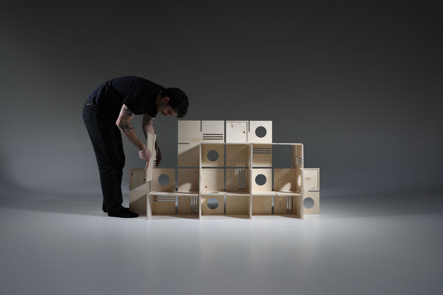 Person assembling a structure made of wooden boxes with circular and rectangular cutouts.