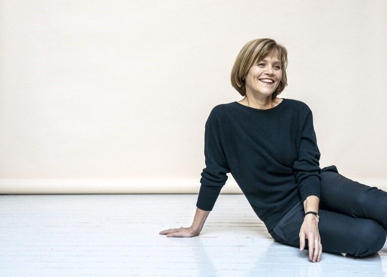 Henkilö, jolla on lyhyet hiukset ja tumma paita sekä housut, istuu vaalealla puulattialla beigeä seinää vasten.