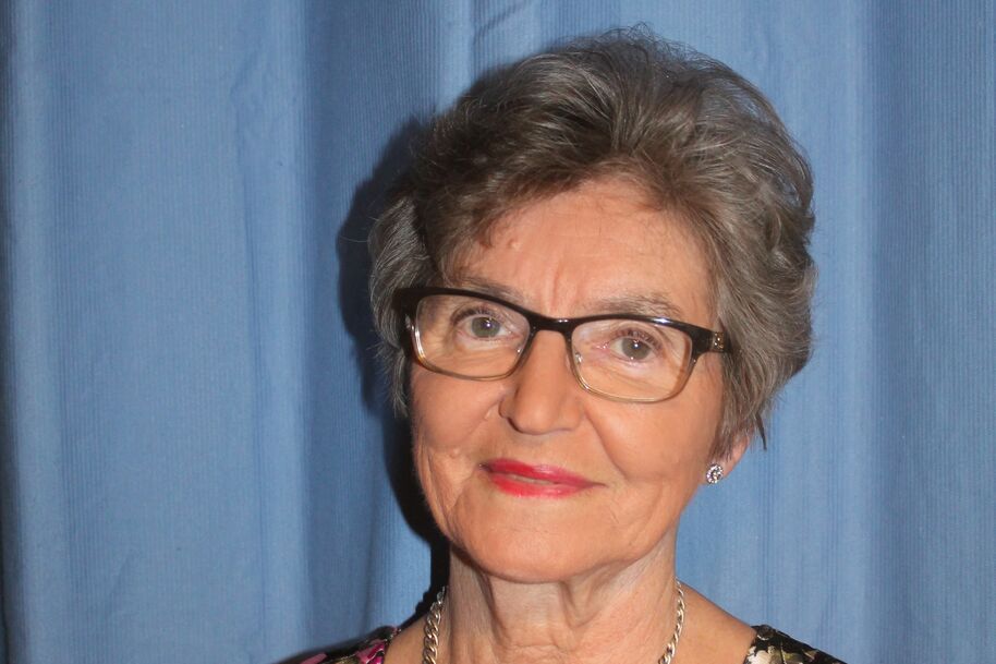 Iäkkäämpi henkilö, jolla on lyhyet harmaat hiukset, kukallinen paita ja kultainen kaulakoru, seisoo sinisen verhon edessä.