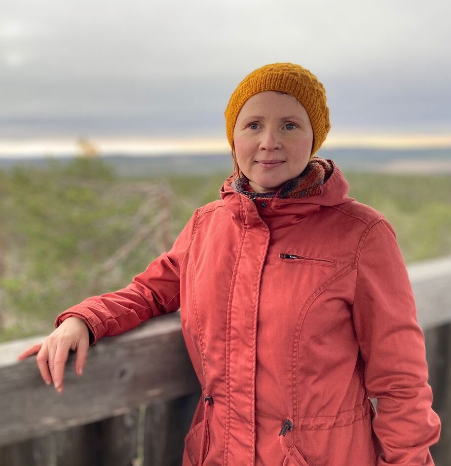 Henkilö punaisessa takissa ja keltaisessa hatussa seisoo puukaiteen vieressä maisemataustalla.