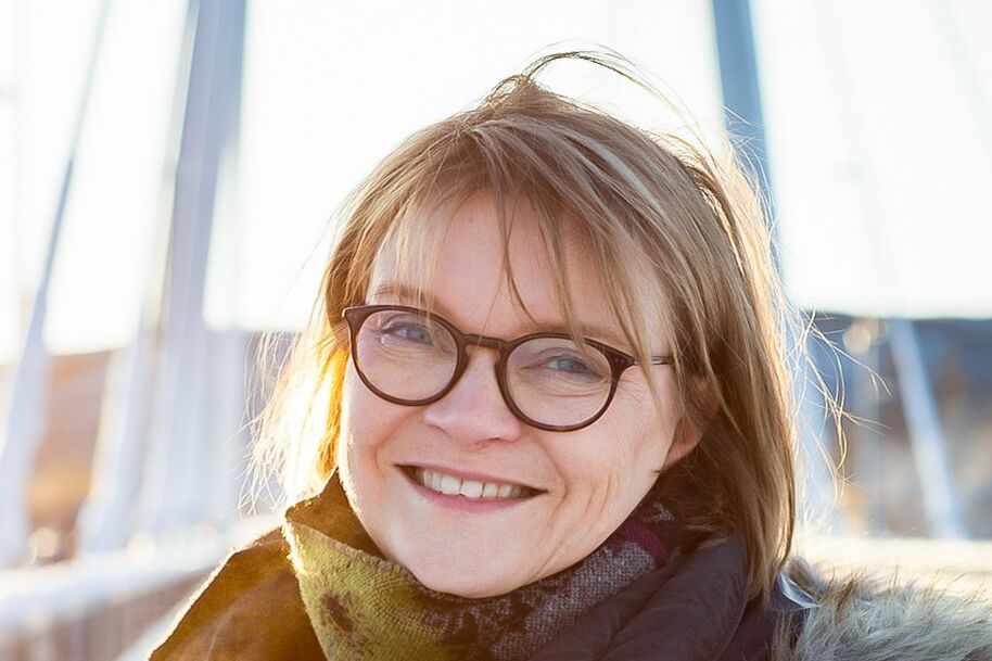 Henkilö, jolla on vaaleat hiukset, huivi ja takki, seisoo ulkona taustalla silta.