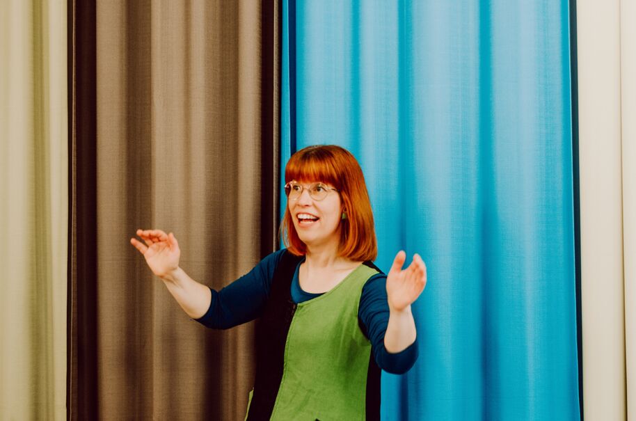 Punatukkainen henkilö vihreässä ja mustassa mekossa seisoo sinisten ja ruskeiden verhojen edessä, kädet ylhäällä.