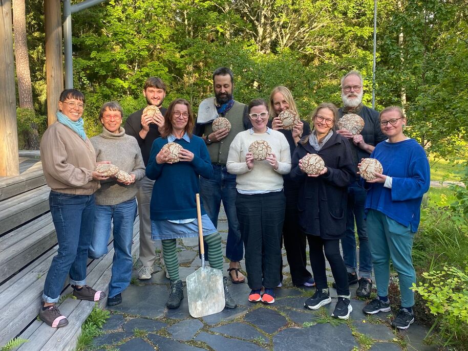 A group of people holding round loaves of bread, standing outdoors on a stone path near wooden steps and greenery.