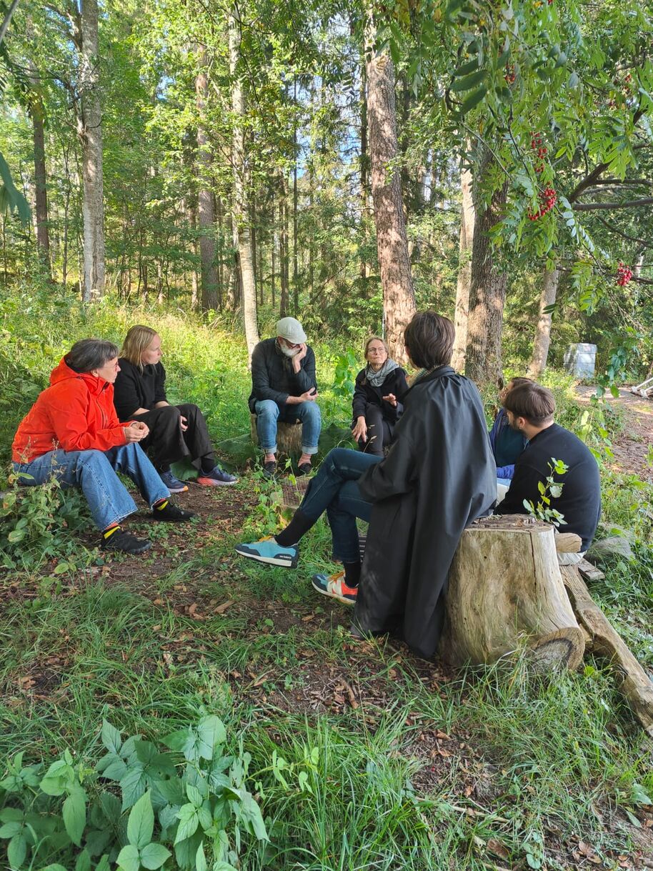 A group of people sitting in a circle outdoors in a forest, some on logs and others on the ground, surrounded by trees and greenery.