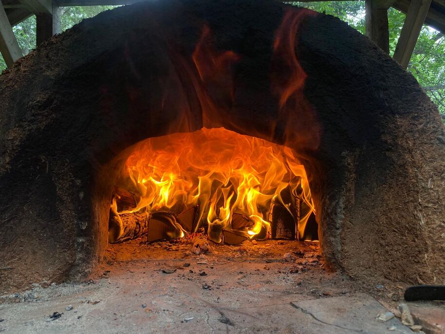 A wood-fired oven with bright flames burning inside. The oven has a rustic, dark exterior and is surrounded by greenery.