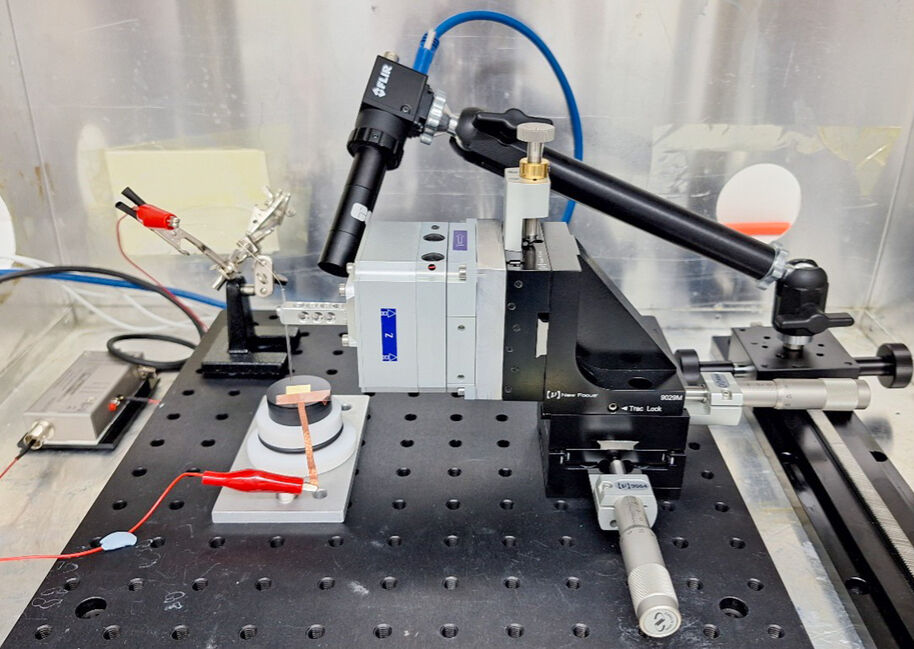 A laboratory setup on a perforated black metal table includes microscopes, clamps, and wires connected to equipment.