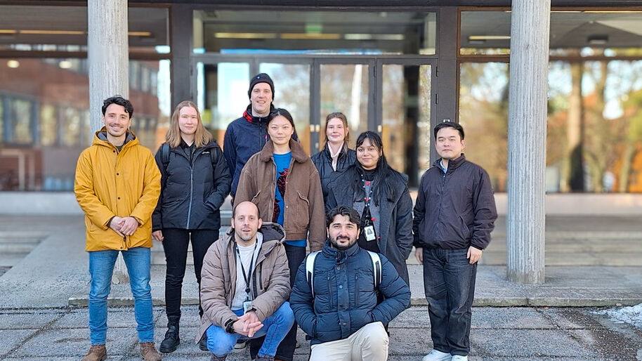 A group of nine people standing outdoors in front of a glass building. They are dressed in winter clothes and are mostly wearing jackets.