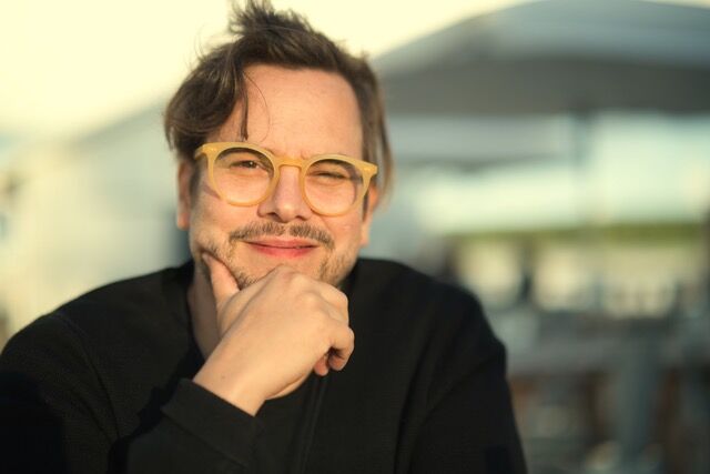 Henkilö ulkona, kasvot sumennettu, musta takki, käsi suun lähellä, tausta kirkas ja pehmeä