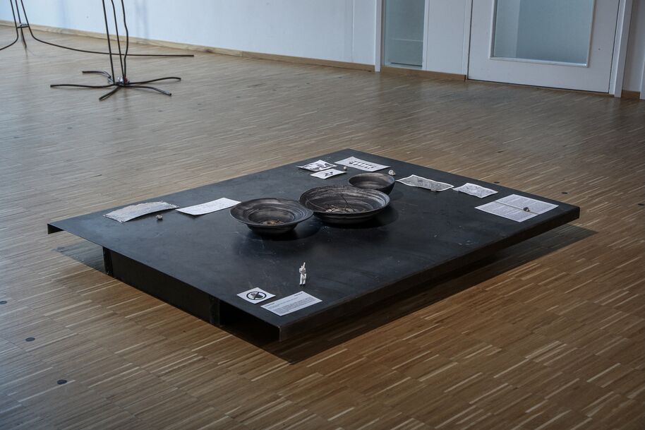Low black platform with metal bowls and scattered papers in a bright wooden-floored gallery