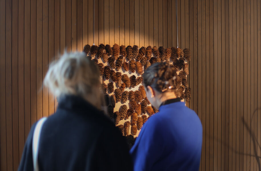 Two people view a wall artwork of pinecones arranged in patterns on vertical wooden panels.