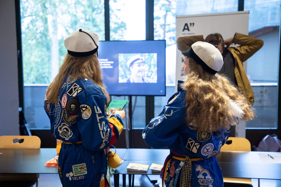 Two students in blue overalls and white caps watch a screen at an indoor event table