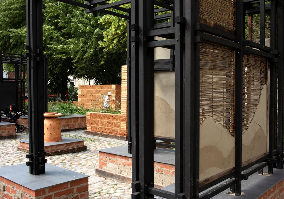 Ulkopaviljonki, mustat rungot, tiilijalustat ja punosseinät, taustalla kävelee hattupäinen lapsi.