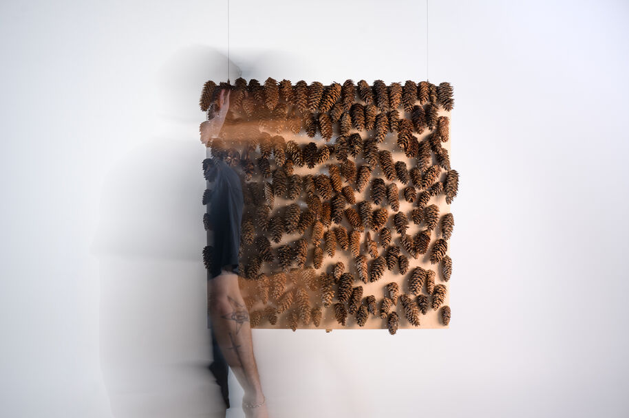 Blurred person passes a beige wall panel densely covered with brown pine cones in a white gallery.