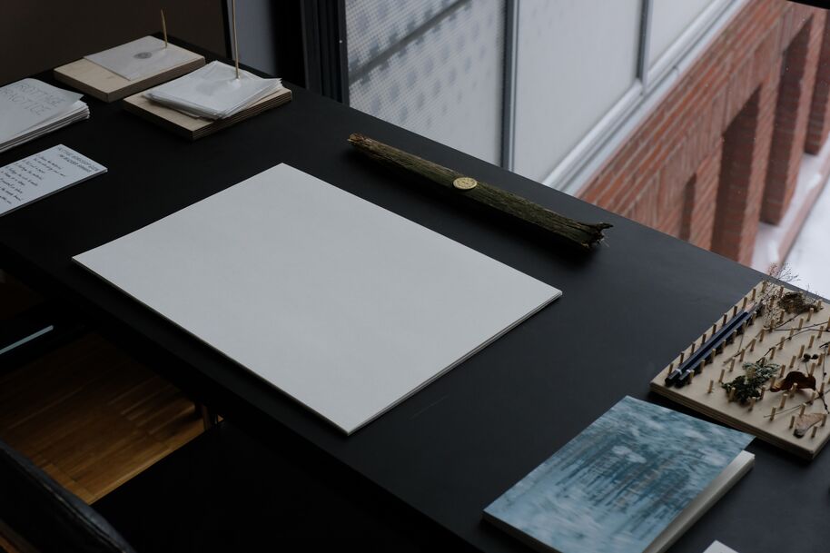Dark desk by a window with blank white sheet, art books and a wooden branch-like object arranged neatly