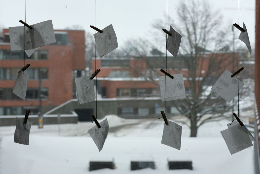 Grey papers hang on strings before a snowy campus with red brick buildings and bare trees outside the window.