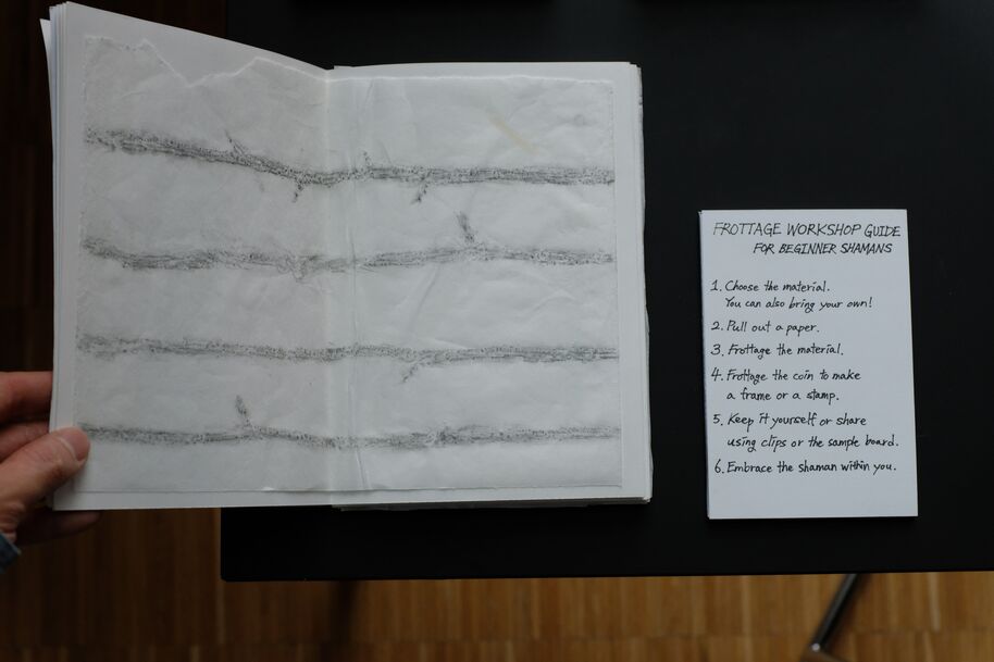 Hand holding sketchbook with grey rubbing lines, beside white frottage workshop guide sheet on dark table