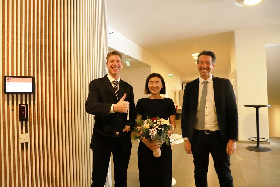 Three dressed-up people indoors, middle person holding a bouquet as man on left gives thumbs-up