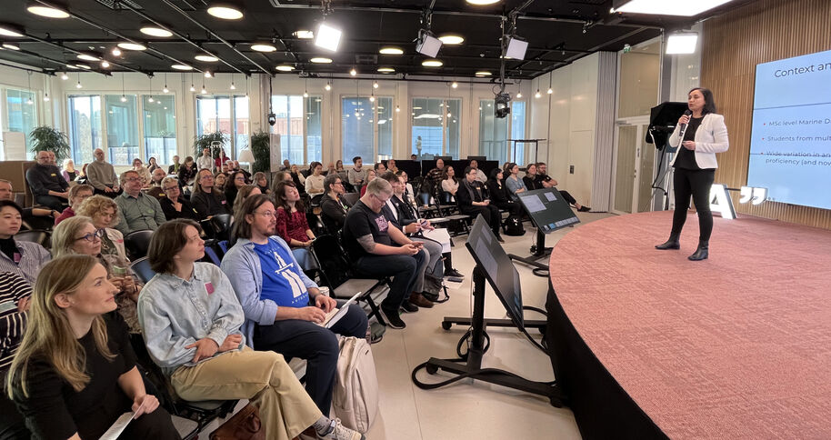 Konferenssisali, yleisö istuu ja kuuntelee pyöreällä lavalla esittävää puhujaa ja näyttöä
