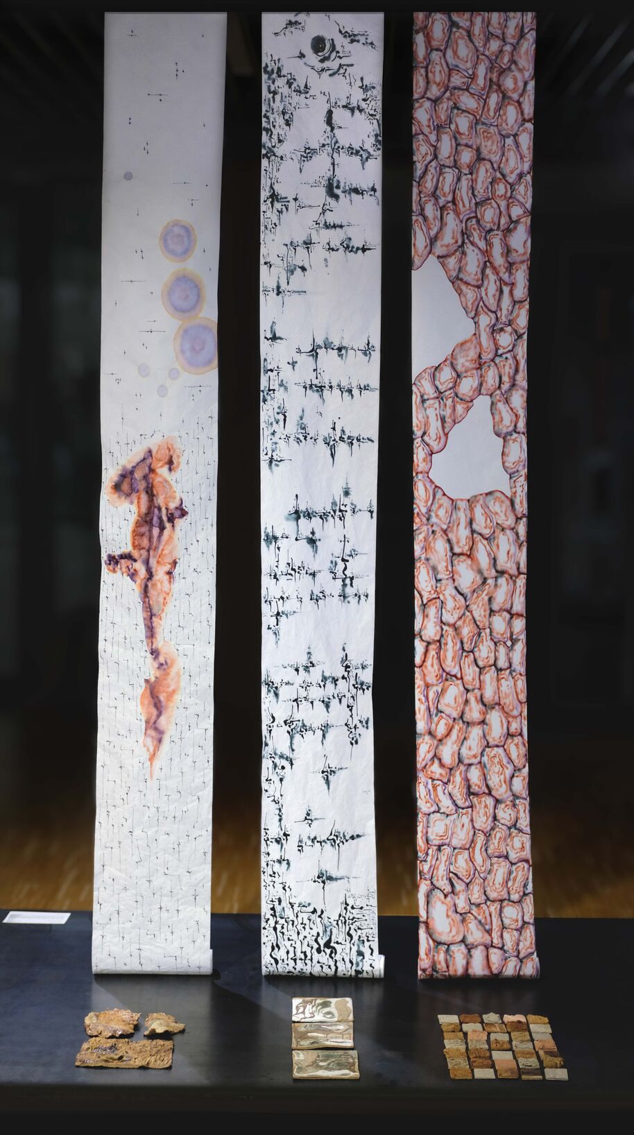 Three hanging fabric panels with organic patterns above small textured tiles on a dark table