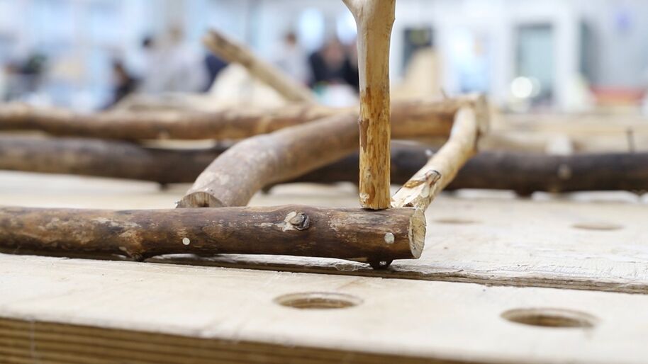 Rough brown wooden branches laid on a pale workbench with round holes in a blurred workshop.