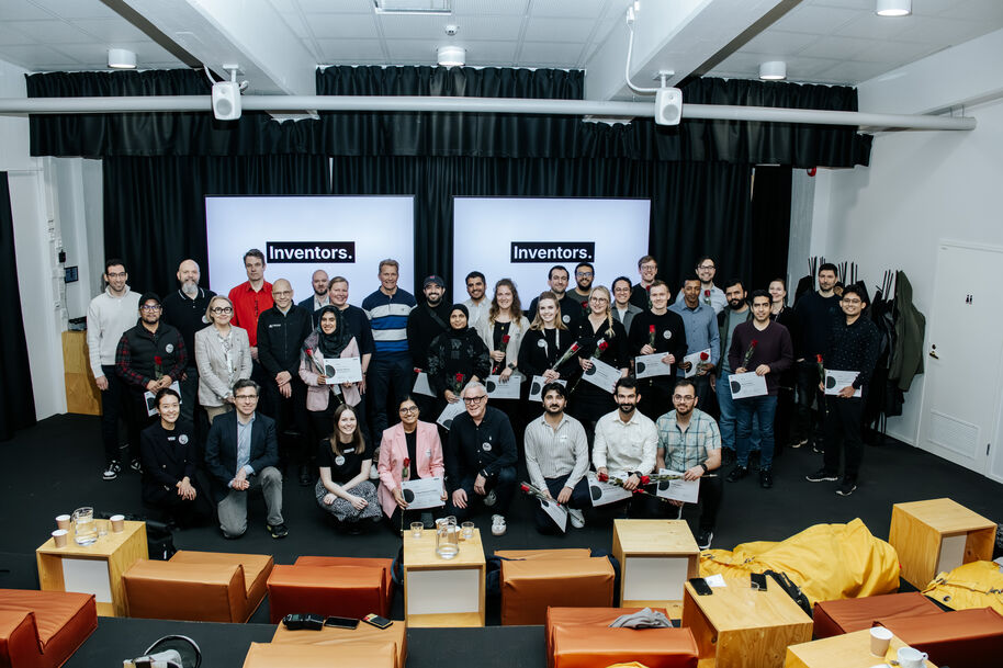 Large group holding certificates and roses in a modern hall, standing before two Inventors screens.