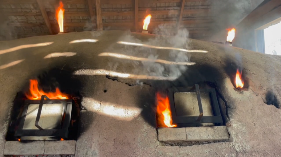 Large clay kiln with orange flames and smoke rising from several square openings indoors