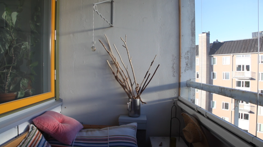 Small glazed balcony with bench, striped cushions and dry branches in a jug, overlooking apartment buildings