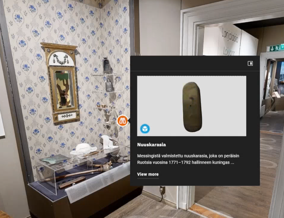 Museohuone, kukkatapetti, peili, vitriini esineineen ja nuuskarasian esittelykortti etualalla.