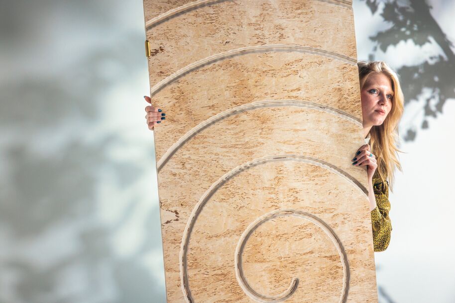 Person peeks from behind tall beige panel with spiral grooves, outdoors in soft light