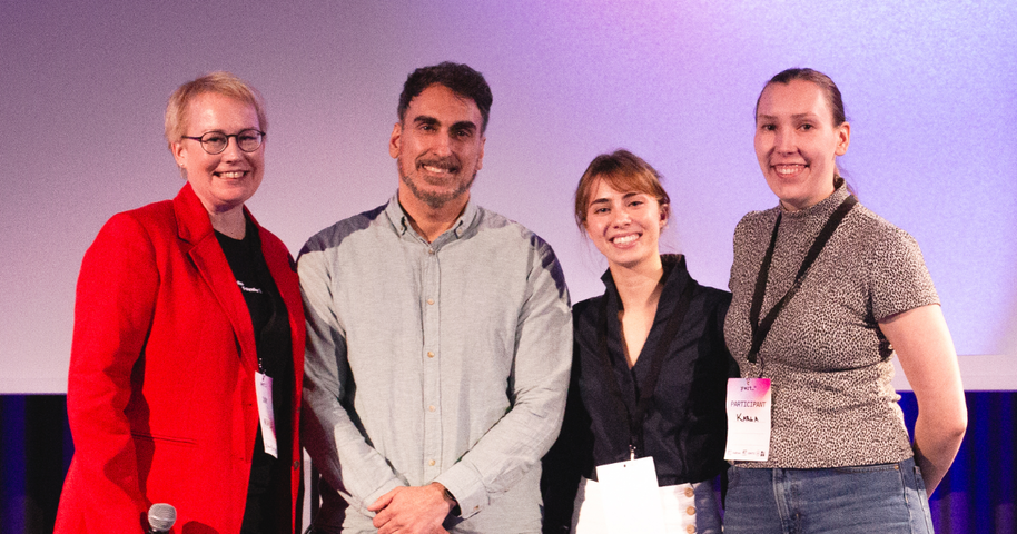 Four smiling people on stage, two with name badges, one in a red blazer, posing under purple lighting