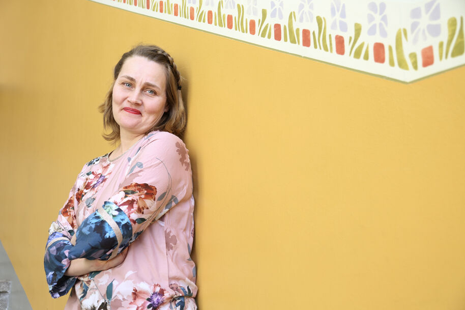 Hymyilevä nainen kukkakuosisessa paidassa nojaa keltaiseen seinään, yläreunassa koristekuvio