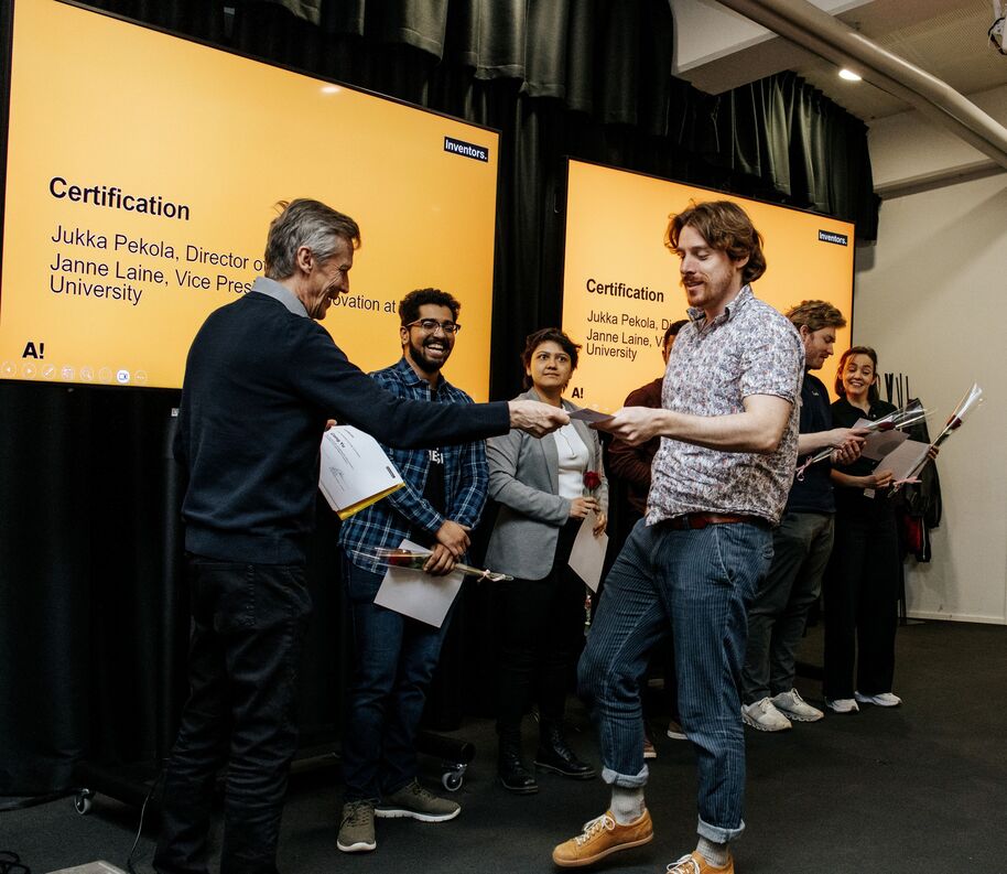 Smiling people receive certificates and roses on stage in front of large yellow presentation screens.