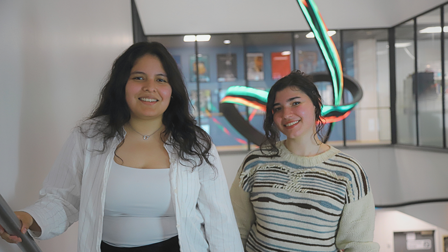 Two smiling young women indoors, one in white shirt, one in striped jumper, with colourful light sculpture behind
