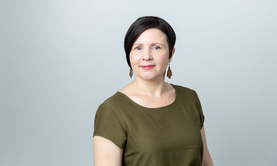 Lyhyttukkainen nainen oliivinvihreässä paidassa ja korvakoruissa vaaleanharmaata taustaa vasten.