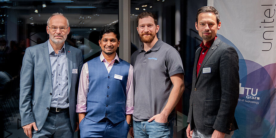 Four men posing at an indoor event, wearing smart clothes in grey, blue and red