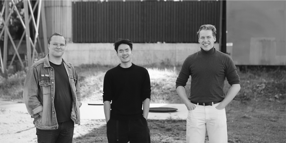 Three smiling men standing outdoors in black and white, hands in pockets, industrial buildings behind