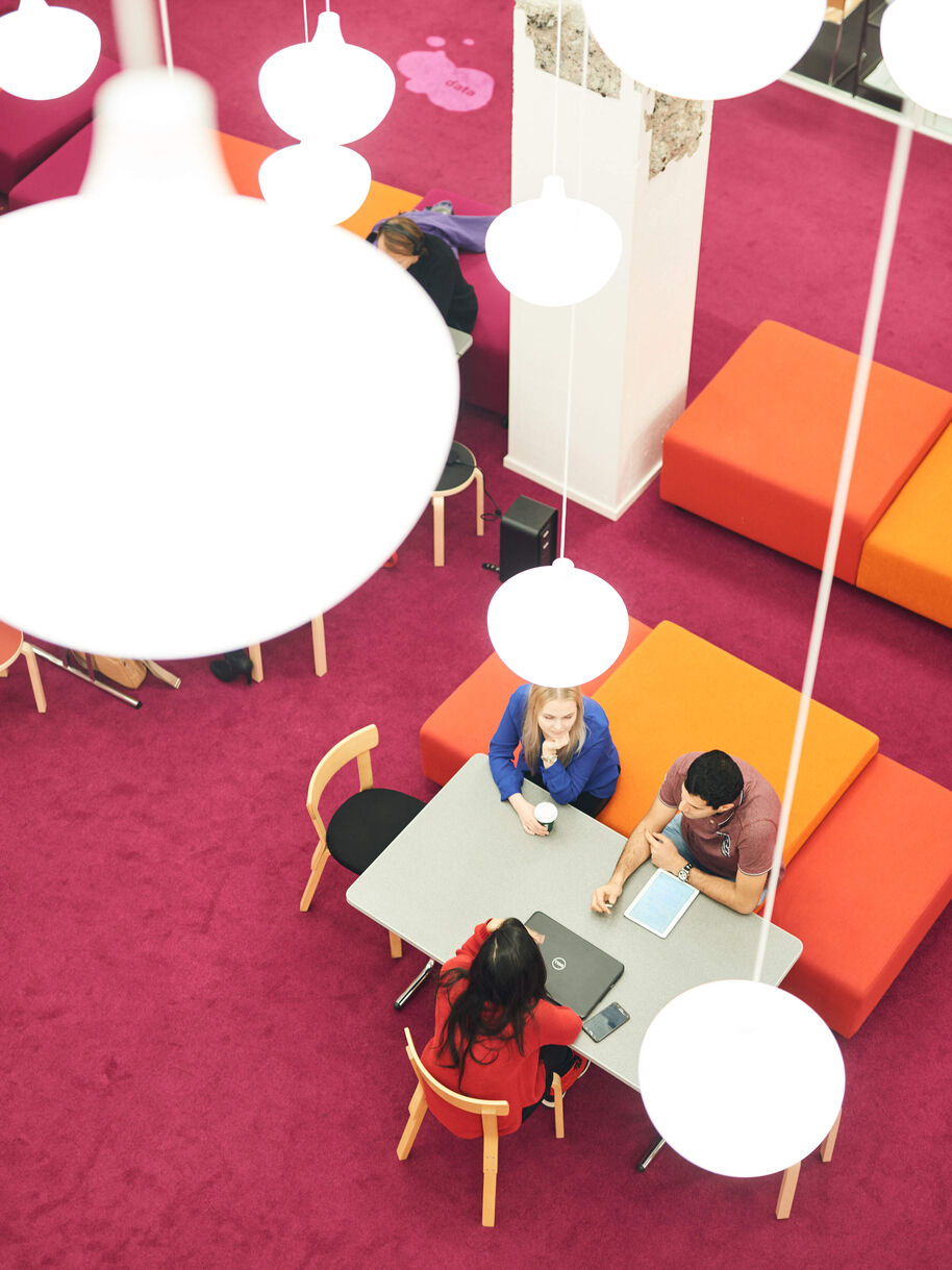Tre personer samtalar vid ett bord i en färgstark lounge med rosa matta, orange soffor och bärbara datorer