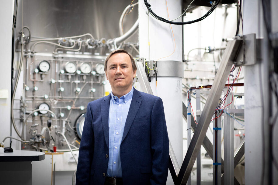 Middle‑aged man in blue blazer stands in industrial lab with metal pipes, gauges and cables behind him