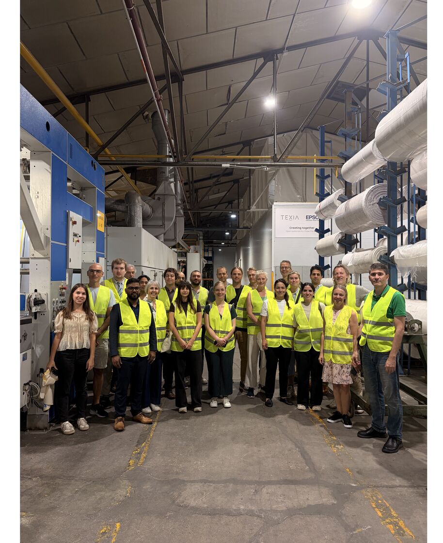 Group of adults in yellow safety vests pose in a textile factory with large fabric rolls and machines.