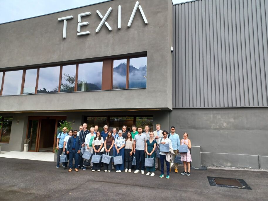 Group of people with gift bags pose outside a grey TEXIA building with large windows and mountain reflections