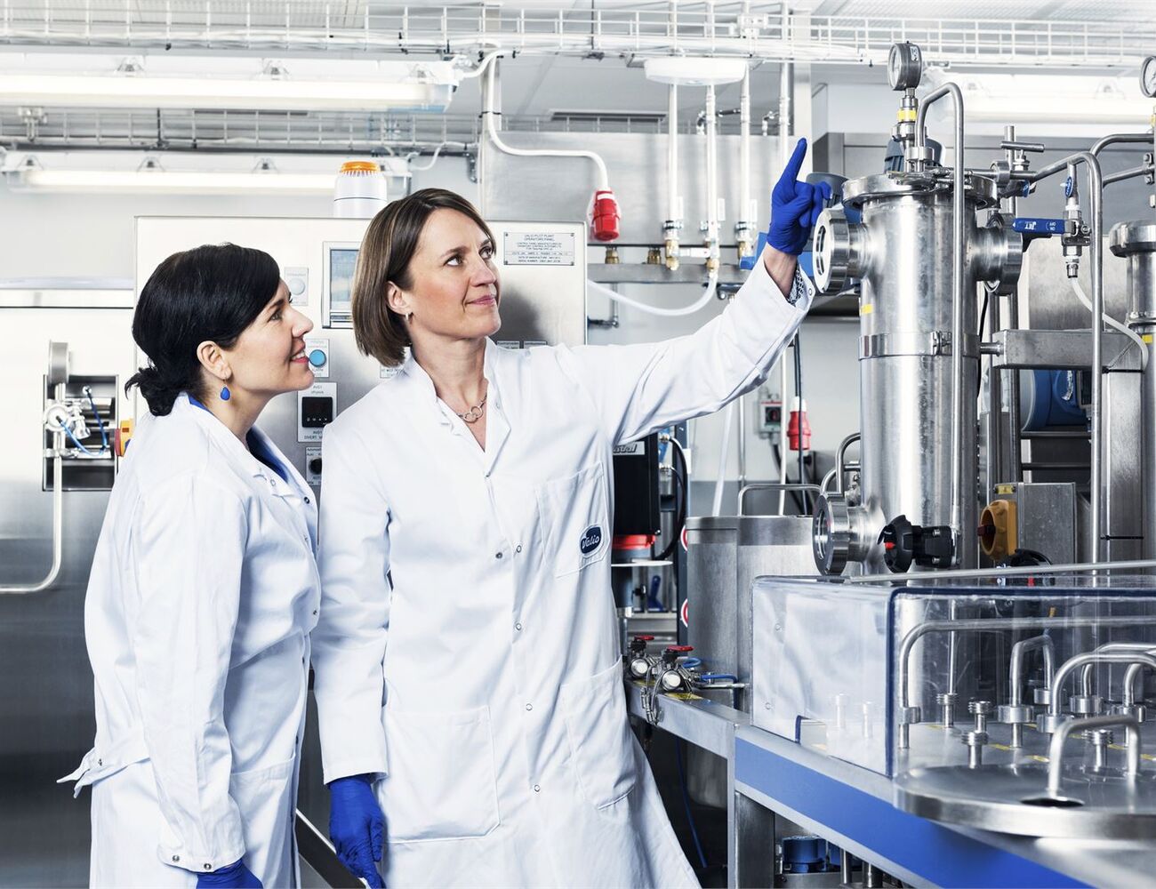 Two lab-coated women in a laboratory looking at pipes.