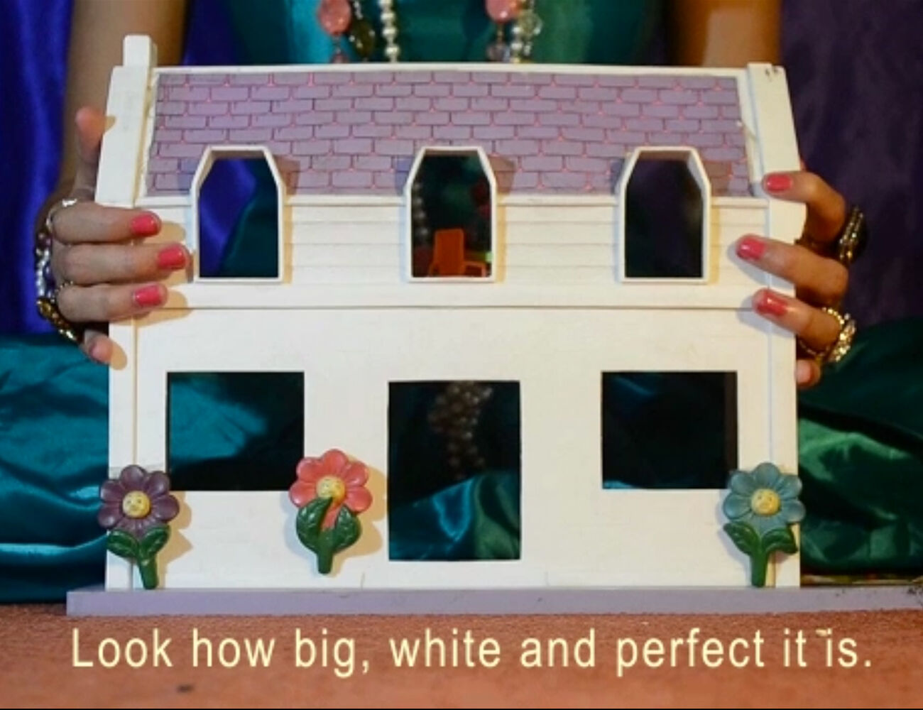 A video still featuring a white doll house with a woman sat behind it