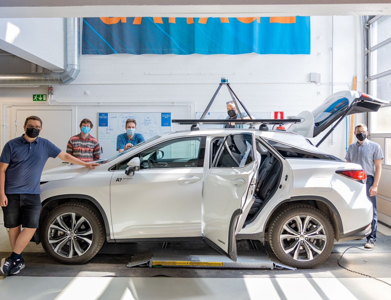 Autonominen A!ex-auto kuvattuna valoisassa autohallissa ympärillään viisi tutkijaa kesäisissä vaatteissa ja kasvoillaan maskit. Valkoisen auton katolla on tekninen laite ja auton tavaratilan kansi on ylhäällä.